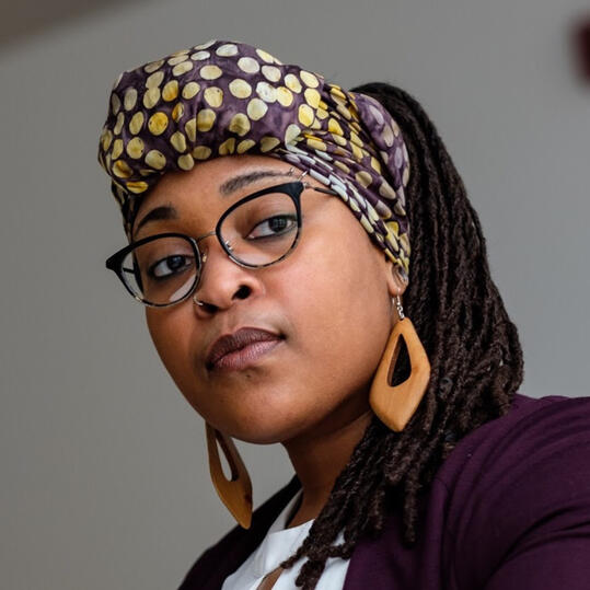 Image: Black queer AFAB person with long locs, a multi-colored headwrap with large light-colored dots, black-framed glasses, and a neutral expression looking off to the right.